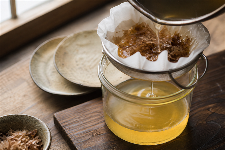 Step 7: pale gold dashi being strained slowly through a paper-lined fine mesh sieve into