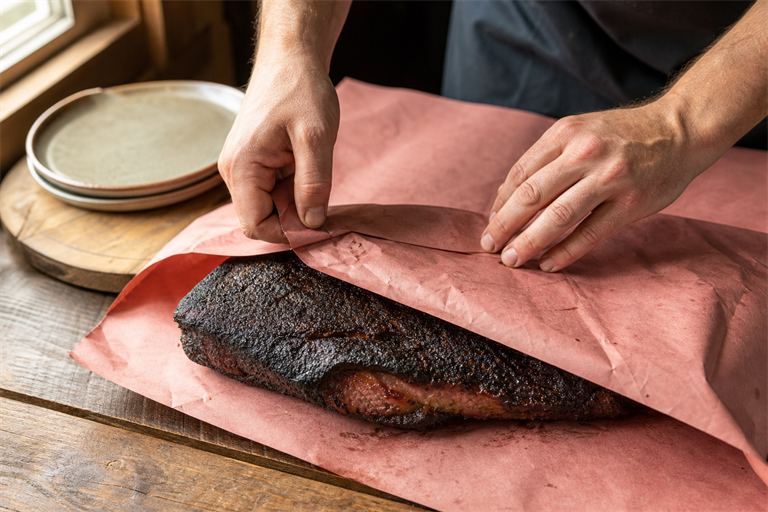 Step 7: a brisket being wrapped in pink butcher paper on a wooden table, dark bark visib