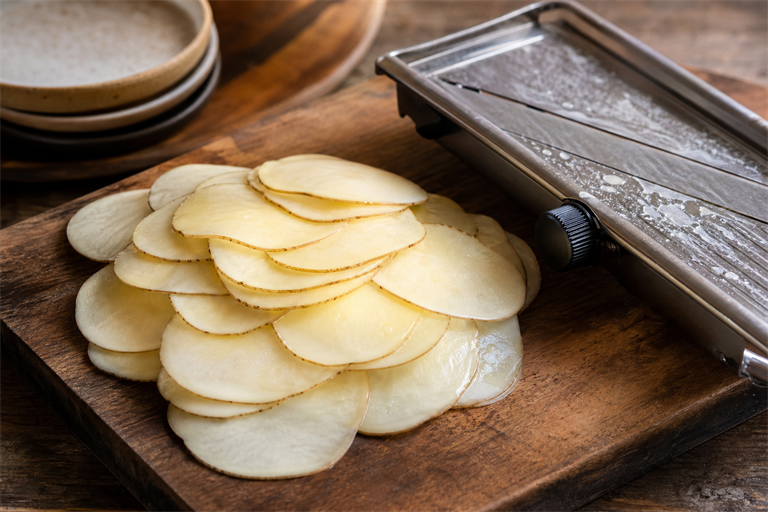 Step 2: thin Charlotte potato slices stacked on a wooden cutting board next to a stainle